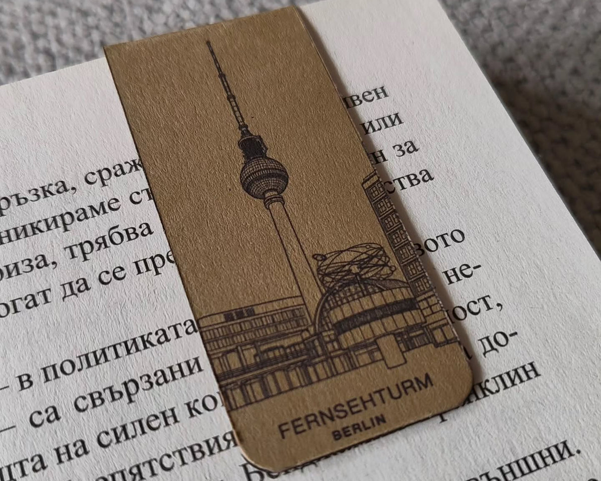 A rectangular kraft paper bookmark with an engraved illustration of the Berlin Fernsehturm (TV Tower) and the text 'FERNSEHTURM BERLIN', resting on an open book with printed text.